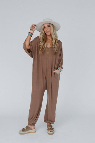 A woman with long blonde hair wears the "Lovely As Ever Harem Jumpsuit" by Three Bird Nest, pairing it with a wide-brimmed hat and accessorizes with bracelets and necklaces. She stands against a plain, light gray background, her left hand in her pocket and her right hand touching her hat.
