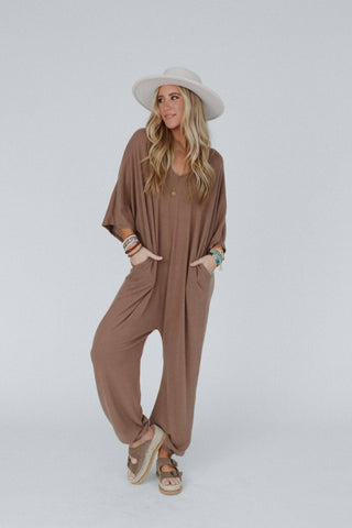 A woman stands in a relaxed pose, dressed in the "Lovely As Ever Harem Jumpsuit" by Three Bird Nest, a boho-inspired, loose brown harem jumpsuit made of comfortable knit fabric, with her hands in the pockets. She accessorizes with a white wide-brimmed hat, multiple bracelets, necklaces, and beige sandals. The background is plain gray.