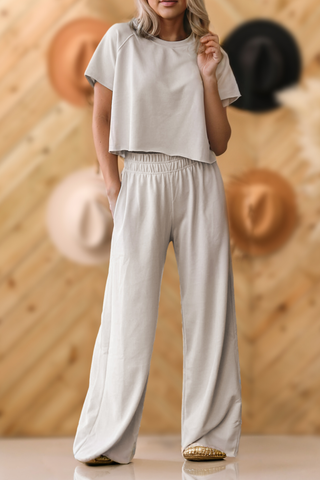 A person wearing the Berkley Women Raw Hem Shirt and Wide Leg Pants Loose Set by Aapparella, in light gray. The outfit features a short-sleeved top and wide-leg pants with hands casually placed in pockets. The casual background displays a wooden wall adorned with several colorful hats, showcasing a perfect setting for daily wear.