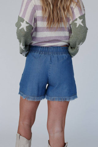 A person with long blonde hair is wearing a sweater with green sleeves featuring white star designs and a striped pattern. They are also sporting the "Meet The Moment Drawstring Shorts" in dark wash from Three Bird Nest, which have a high-rise fit and frayed hem. The individual is facing away from the camera, wearing light-colored boots.