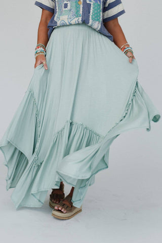 A person is wearing the Logan Forever Beauty Maxi Skirt in pale sage by Three Bird Nest, featuring a high-rise elastic waistband and ruffled boho design. They are paired with a blue and white patterned top, colorful bracelets on their hands, and brown sandals. Against a plain white backdrop, they hold up the sides of the skirt.
