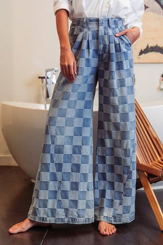 A person stands on a wooden floor wearing the Wash Two denim Checkered Jeans from Western Edge Boutique. These high-waisted, wide-leg jeans feature a stylish blue checkered pattern and pleated front. A white shirt is partially visible, enhancing the chic ensemble. In the background, a freestanding bathtub and a wooden chair contribute to the serene ambiance.