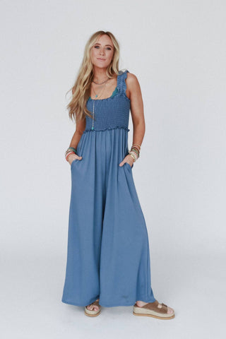 A woman stands against a plain background wearing the "Nothing Compares Smocked Ruffle Jumpsuit - Denim Blue" by Three Bird Nest. The comfortable, lightweight blue jumpsuit is sleeveless and features flowy pant legs and ruffle straps. With long blonde hair, she accessorizes with multiple bracelets, necklaces, and tan platform sandals. Her hands rest in the jumpsuit pockets.