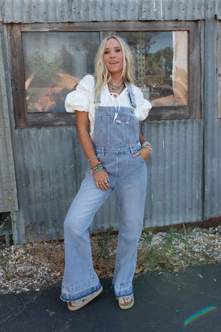 A woman with blonde hair stands confidently in front of a corrugated metal wall, embodying a boho vibe. She is wearing comfortable fabric in light blue Cali Dream Denim Overalls - Light Wash by Three Bird Nest, paired with a white puff-sleeve blouse and tan sandals. She accessorizes with bracelets and necklaces. The window behind her reflects blurred outdoor scenes.