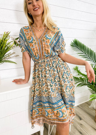A person in the vibrant Finn Tie Back High Low Dress by Anna Kaci Wholesale stands in a room with white paneled walls. The dress showcases blue and orange floral designs. They are smiling with their blonde hair down, while green plants adorn a white table and the background.