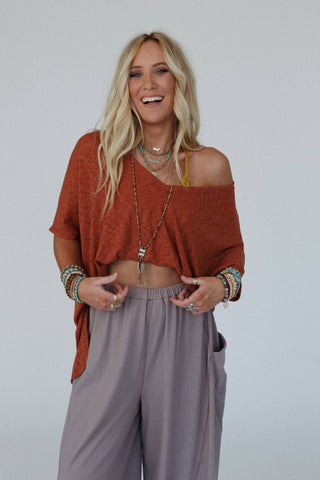 A blonde woman smiles while wearing a cayenne Kingsley Warm Nights V Neck Top from Three Bird Nest and light brown loose pants. She accessorizes with layered necklaces, various bracelets, and rings. Her relaxed and stylish outfit, made of comfortable ribbed knit fabric, exudes a casual, bohemian vibe with a touch of western style.
