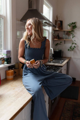 A person with long blonde hair sits on a kitchen counter, holding a small cup and smiling. They are wearing the Three Bird Nest Nothing Compares Smocked Ruffle Jumpsuit in Denim Blue and various bracelets. The kitchen, featuring wooden countertops, white tiles, plants, and natural light coming through the window, showcases a warm ambiance.