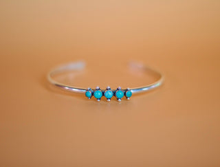 The Turquoise & Sterling Silver Cluster Cuff Bracelet 925 by Moxie Maine features five small turquoise gemstones evenly spaced along the center of a delicate sterling silver bracelet. Set against a smooth, warm beige background, this design highlights its simple and elegant charm.