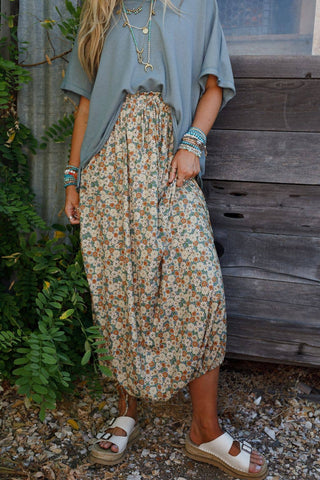 A person with long hair is wearing a loose blue-gray T-shirt, the Solstice Song Cinched Skirt - Khaki by Three Bird Nest, and white sandals. They are accessorized with layered necklaces, bracelets, and rings. The background features greenery and a wooden surface.