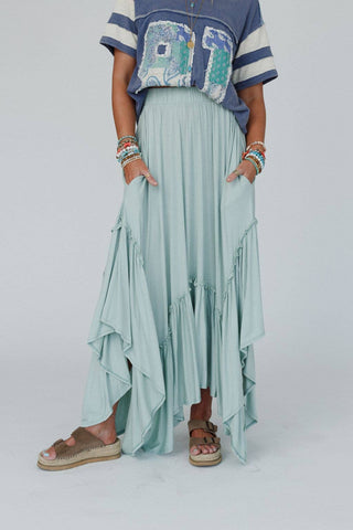 A person is wearing a blue graphic T-shirt and the Logan Forever Beauty Maxi Skirt in Pale Sage from Three Bird Nest, which features a high-rise elastic waistband. They have their hands in the skirt pockets and are accessorized with layered bracelets on both wrists. They are also wearing beige platform sandals.