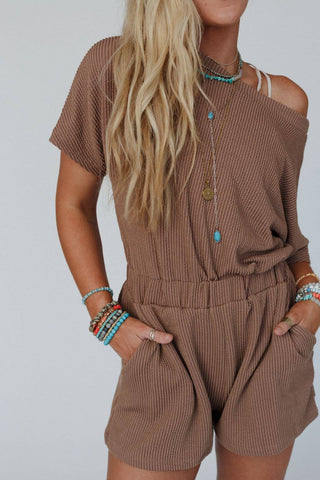 A person with long, wavy blond hair wearing a brown Covey Romper - Camel by Three Bird Nest poses against a plain background. The short-sleeve romper features pockets, ribbed knit fabric, and an elastic waistband. They accessorize with layered necklaces, bracelets, and rings, some showcasing turquoise stones.
