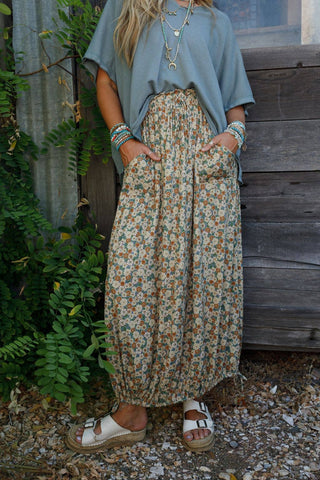 A person wearing a loose, light blue top, the Solstice Song Cinched Skirt in khaki by Three Bird Nest, multiple necklaces, and bracelets stands against a wooden wall with green foliage nearby. They have their hands in their skirt pockets and are wearing beige sandals.