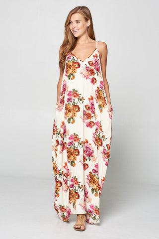 A woman with long, light brown hair wears the VANILIA BAY Ava Floral Summer Dress Curvy, a flowing white maxi dress adorned with bright floral patterns. She is standing against a plain, light gray background with her hands in the dress's side pockets, smiling and looking slightly to her left.