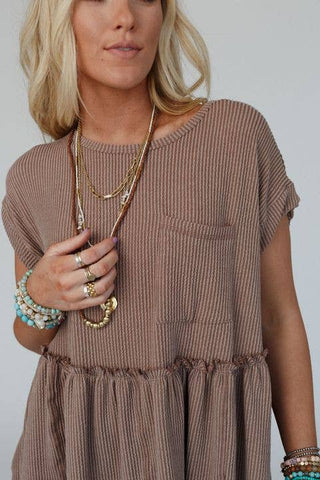 A person with long, blonde hair is wearing a loose-fitting, short-sleeved Meadow Subtle Escape Textured Dress in brown ribbed fabric from Three Bird Nest, featuring a pocket on the chest. They are accessorized with layered necklaces and multiple bracelets in turquoise and gold tones, giving off boho western vibes. The background is neutral.