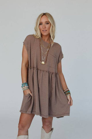 A woman with blonde hair is wearing a Meadow Subtle Escape Textured Dress by Three Bird Nest. The dress is loose, short-sleeved, and made from light brown ribbed fabric. She accessorizes with layered necklaces and bracelets on both wrists, and completes her look with white knee-high boots that add boho western vibes. She poses effortlessly with one hand in her pocket.