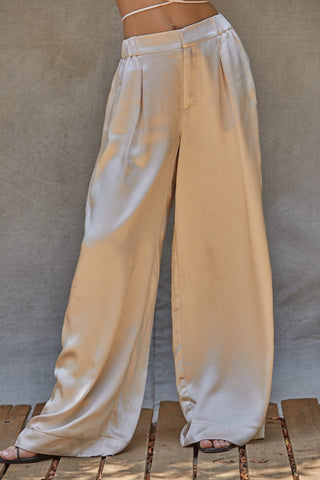 A person wearing high-waisted, wide-leg, beige Trinity Satin Stunner Pants by By Together poses against a neutral backdrop. The glossy satin pants feature pleats and a slightly loose fit. The person's upper body is not visible, emphasizing the pants' style and fabric texture.