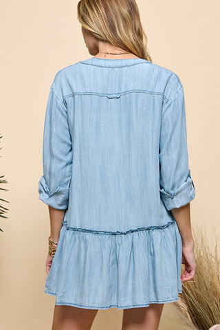 A person wearing a Billie Denim Blue Button V Neck Ruffle Dress from Western Edge Boutique is facing away. This dress, made from Tencel, showcases a loose ruffled hem and has rolled-up sleeves. The background features a soft beige tone with greenery on the side.