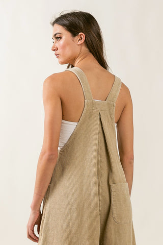 A woman with long dark hair is seen from the back, wearing the Briley Corduroy Overalls Wide Leg by BiBi in beige over a white top. The overalls feature wide straps that cross at the back and a pocket at the bottom, offering a comfy fit reminiscent of classic styles. She is standing against a plain, light-colored background.