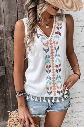 A person wearing the Western Edge Boutique's Redner Sleeveless V Neck Embroidery Tassel Hem Top, paired with blue denim shorts. They are accessorized with layered necklaces, bracelets, and a wide-brimmed hat, standing against a rustic wooden and white brick background.