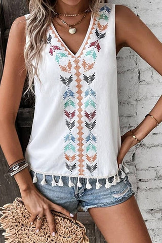 A person in denim shorts and the Redner Sleeveless V Neck Embroidery Tassel Hem Top from Western Edge Boutique stands near a white brick wall. They are holding a straw clutch and wearing bracelets and a necklace.