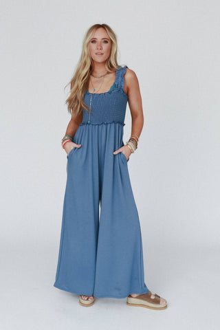 A woman with long blonde hair stands against a plain white background, wearing the Nothing Compares Smocked Ruffle Jumpsuit by Three Bird Nest in a lightweight stretch fabric. The sleeveless denim blue jumpsuit features wide pant legs and ruffled straps, with her hands casually tucked into the pockets. She accessorizes with various necklaces and bracelets, completing her look with sandals.