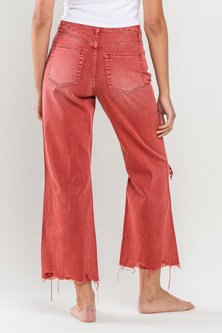 A person wearing a white top and VERVET by Flying Monkey Ashlyn Vintage Distressed Crop Flare Leg in a vibrant red, featuring loose-fitting, high-waisted distressed hems and frayed edges. The photo is taken from the back, showcasing the rear pockets and the casual, relaxed fit of the high-rise jeans. The person is barefoot.