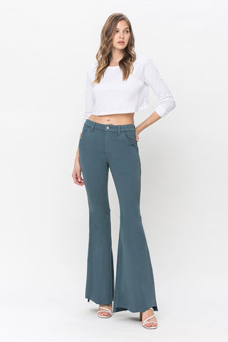 A person with long hair is standing against a plain background. They are wearing a white long-sleeve crop top, Delaney Super High Rise Wide Leg Jeans by VERVET by Flying Monkey, and white heeled sandals. Their hands are in their pockets as they look toward the camera.