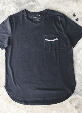 A black short-sleeved Jillian "Handy as a Pocket" Tuxedo Tee by 2 Fly, a true closet staple, lies flat on a concrete surface. It features a small chest pocket with a minimalist white arrow design pointing horizontally. The shirt has a rounded hem and a crew neck. The tag is visible at the back of the neck.