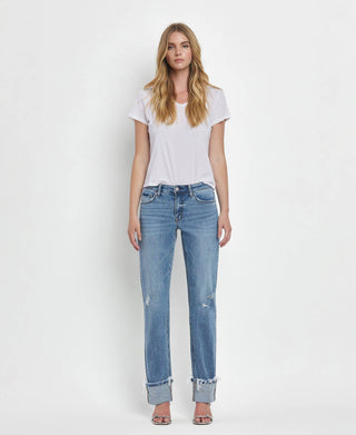 A woman stands facing forward against a white background, wearing a white t-shirt and Dezie Mid Rise Cuffed Straight Jeans by Lovervet by VERVET. Her jeans feature soft stretch denim and rolled cuffs. She has long, loose hair and is wearing open-toed shoes.