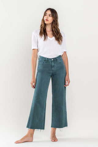 A woman with long, wavy hair wearing a white short-sleeve V-neck shirt and teal VERVET by Flying Monkey Bexlee High Rise Crop Wide Leg Jeans is standing barefoot against a plain light background. She is looking slightly to the side, and her arms are relaxed by her sides.