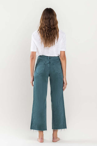 A person with long, wavy hair stands facing away from the camera, wearing a white short-sleeve shirt and Bexlee High Rise Crop Wide Leg Jeans in teal with frayed hems by VERVET by Flying Monkey. The background is a plain, light-colored wall.