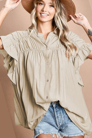 A person with wavy blonde hair smiles while wearing the silky woven "Livey Loose Fit Silky Cappuccino" blouse by BiBi, complete with ruffled sleeves and a straw hat. They are also donning light denim shorts, revealing visible wrist tattoos against a solid neutral background.