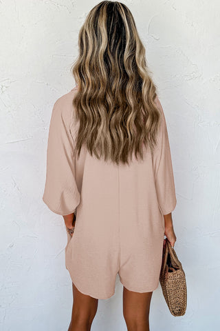 A woman with wavy, blonde hair stands with her back to the camera, showcasing the Brinley Apricot Half Button Collared Loose Romper from Western Edge Boutique. She is holding a woven straw handbag against a plain, light-colored textured wall. This casual romper for women features an oversized silhouette, perfect for effortless style.