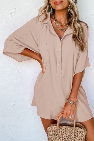 A woman wearing the Brinley Apricot Half Button Collared Loose Romper from Western Edge Boutique poses against a white textured wall. She accessorizes with layered necklaces and bracelets, carrying a woven straw handbag. Her wavy blonde hair partially covers her face, creating an effortlessly chic look reminiscent of classic summer style.