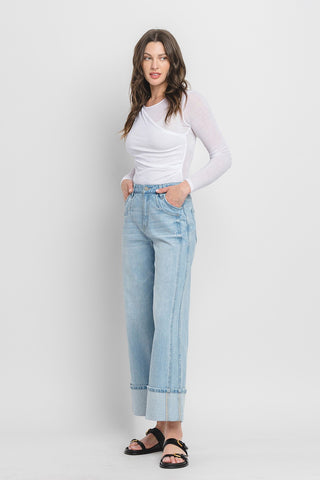 A woman wearing a long-sleeve white top and the Hazen Super High Rise Cuffed Baggy Wide Leg jeans from Western Edge Boutique stands against a plain white background. Her hands are in her pockets, and she is wearing black sandals.