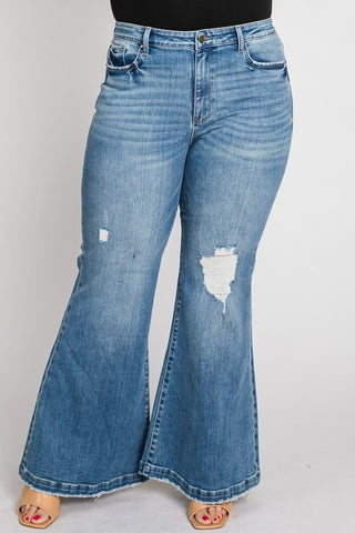 A person is wearing Petra153's Cypress PLUS - Super High Rise 70s Flare Jeans in light blue, featuring flared legs and two distressed patches on the right thigh. The super high-rise jeans with front pockets are paired with open-toe shoes displaying painted toenails against a plain white background.