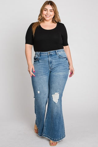 A woman with long, light brown hair smiles while wearing a black short-sleeve top and the Cypress PLUS - Super High Rise 70s Flare Jeans by Petra153, featuring distressed detailing at the knee. She stands against a plain white background, exuding confidence and comfort in her outfit.