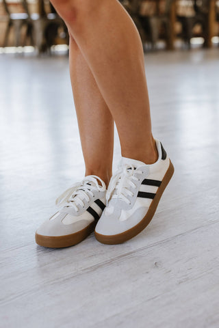 Close-up of a person in Ness Striped Lace Up Flat Sneakers by Liam & Company, featuring black stripes and gum soles on light wooden flooring. The sneakers add classic style against the softly blurred backdrop, focusing on their trendy flair with only the legs visible.