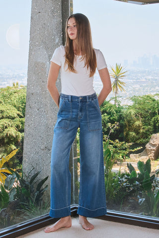 A woman with long hair stands barefoot indoors, dressed in a white T-shirt and the Radley High Double Cuffed Baggy Wide Leg jeans from Western Edge Boutique. She's near large windows overlooking lush greenery and a distant cityscape, relishing in the stylish comfort and ease offered by these jeans.