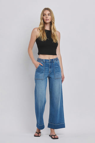 A woman stands against a light background wearing a black sleeveless top and the Radley High Double Cuffed Baggy Wide Leg jeans from Western Edge Boutique. She has long, wavy blonde hair and poses with one hand in her pocket. Embracing comfort stretch denim, she completes her look with stylish black sandals.