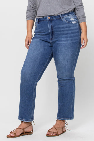 A person models a gray long-sleeve shirt paired with the Coop Curvy High Rise Stretch Slim Straight jeans from Western Edge Boutique. These blue distressed jeans have a high waist and frayed hem, enhancing their style with comfort stretch denim. The stylish ensemble is completed with brown strappy sandals as they pose against a plain background.