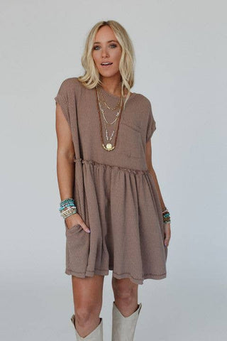 A woman with long, wavy blonde hair is wearing the Meadow Subtle Escape Textured Dress from Three Bird Nest. The dress features a brown, short-sleeved design made from ribbed fabric. She has layered necklaces and bracelets on both wrists and stands with her hands in her dress pockets, exuding effortless boho western vibes against a plain white backdrop.