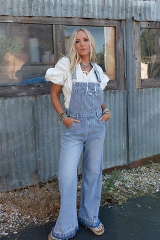 A woman with long blonde hair stands in front of a corrugated metal building, showcasing the comfortable Three Bird Nest Cali Dream Denim Overalls in Light Wash, paired with a white puff-sleeve blouse and boho bracelets on both wrists. Her hands rest casually in her overalls' pockets as she looks towards the camera.