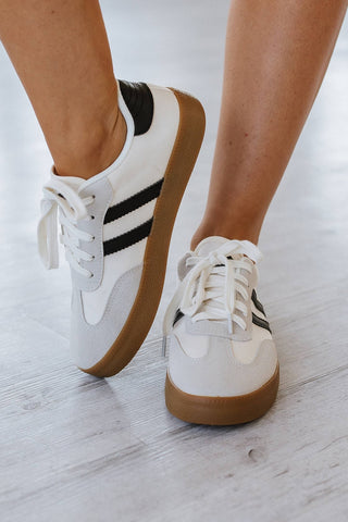Close-up of a person wearing Ness Striped Lace Up Flat Sneakers by Liam & Company. The sneakers have black stripes, white laces, and gum soles on a light floor. One foot is raised to highlight the sneaker's sleek side.