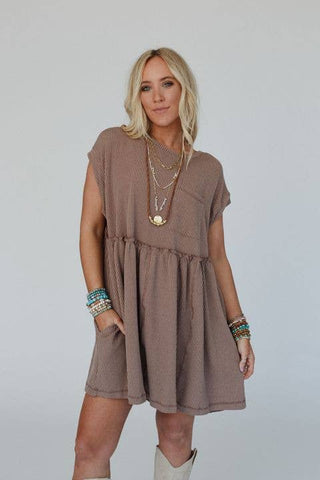 A person with long blonde hair is wearing the Meadow Subtle Escape Textured Dress by Three Bird Nest. This loose, short-sleeved dress features a brown color with white vertical stripes, perfect for portraying boho western vibes. They accessorize with layered necklaces and colorful bracelets. Standing in front of a plain white background, their hands rest comfortably in the dress pockets.
