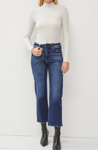 A person is wearing a light grey, long-sleeve, high-neck sweater tucked into dark blue 0live High Rise Wide Leg Crop Jeans non distressed by Petra153. They are also wearing black ankle boots. The flattering fit is perfect for petite figures as they stand against a plain white background with one hand in their pocket.