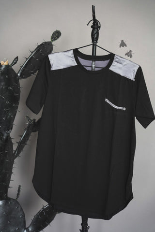 The Blaken Approved Tee from 2 Fly, a black short-sleeve shirt with gray shoulder panels and a small horizontal gray zippered pocket on the left chest, is hung on a black hanger. Featuring new cotton fabric, the backdrop showcases a large cactus with dark green spines, and two drawn flies appear to the right of the shirt.