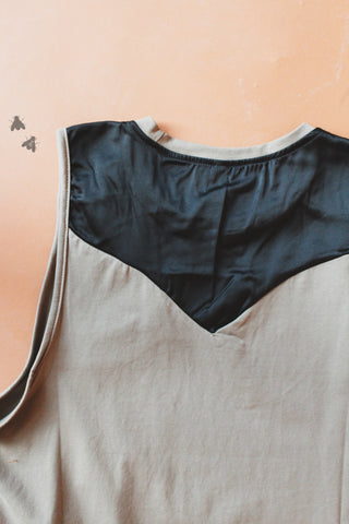 The Tanks to Wanette Tank by 2 Fly is laid out flat, showcasing its light brown lux fabric with a black triangular neckline detail at the back. Two small flies are seen on the upper left side of the image, which is set against a plain brown background.