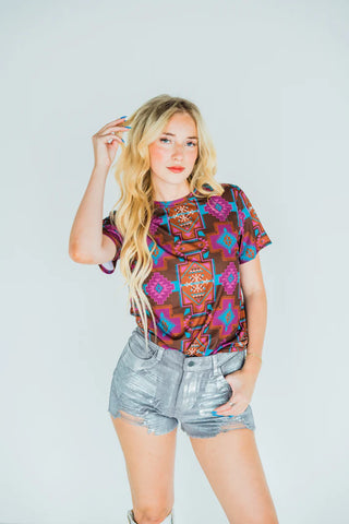A blonde woman with wavy hair is posing against a plain backdrop, confidently wearing 2 Fly's COLOR ME CHROME ensemble—a vibrant, geometric-patterned shirt paired with chic silver chrome shorts. Her left hand is raised near her face, exuding confidence.
