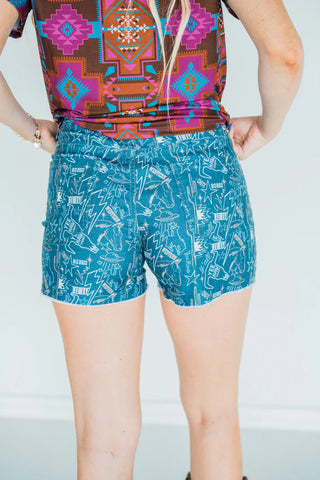 A person wearing the "Call her Cosmo" top from Western Edge Boutique and a pair of super stretchy denim shorts featuring white abstract designs stands with their back to the camera.